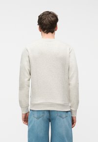 Άνδρας με κοντά κ αστανά μαλλιά που φοράει ανοιχτό γκρι φούτερ και μπλε τζιν, γυρισμένος πλάτη στην κάμερα μπροστά σε λευκό φόντο.