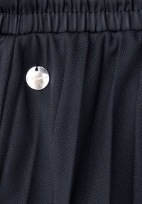 Černá síťovaná látka s nařaseným elastickým pasem a malou kulatou stříbrnou cedulkou připevněnou poblíž horního okraje.