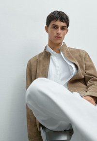 Beige Wildlederjacke mit hohem Kragen, getragen über einem weißen Hemd mit Knöpfen. Die Person hat kurzes, dunkles Haar und sitzt auf einem Metallhocker.