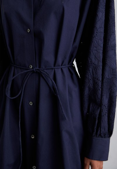 Granatowa sukienka z zapięciem na guziki z przodu, wiązanym w pasie oraz teksturowanymi koronkowymi rękawami zdobionymi kwiatowymi wzorami i detalami w formie oczek. Gładka powierzchnia materiału.