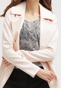 Světle růžový dlouhý kabát s výřezovým límcem, nošený přes černo-bílé tričko s paisley vzorem. Materiál působí měkce a má texturu.