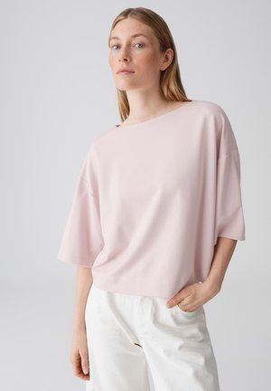Vrouw met steil blond haar draagt een los, lichtroze top met korte mouwen en witte broek, staand tegen een effen lichtgrijze achtergrond.