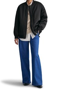 Veste bomber noire avec poignets côtelés, portée sur un pull léger et une chemise blanche, associée à un jean denim bleu à jambes larges et des chaussures noires.