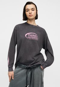 Νεαρό άτομο με κοντά σκούρα μαλλιά που φορά μια ξεθωριασμένη μαύρη μακρυμάνικη μπλούζα με ροζ γράμματα "THINK DIFFERENT" και γκρι παντελόνι με κορδόνι.