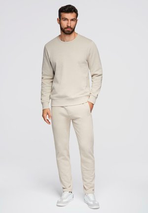 Uomo con barba che indossa una felpa beige, pantaloni della tuta abbinati e scarpe da ginnastica bianche in piedi davanti a uno sfondo chiaro e uniforme.