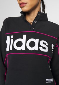 Svart sweatshirt med hög krage, tryckknappar och en djärv vit "adidas"-logo. Har rosa accentstygning och en liten varumärkesetikett vid fållen.