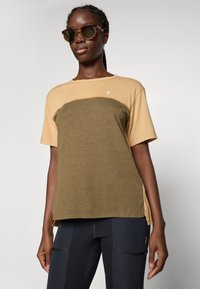 Kvinne iført en tofarget beige og olivengrønn kortermet skjorte, sorte leggings med glidelåslommer, og solbriller, mot en ensfarget lys bakgrunn.