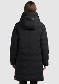 Melns pūkains mētelis ar kapuci, ar spīdīgu logotipa uzlīmi. Mētelis ir garš, tai ir kvadrātveida struktūra un sānu rāvējslēdzēja kabatas.