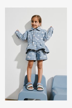 Set de jachetă și pantaloni scurți din denim, culoare albastru deschis, cu modele albe în formă de inimă. Jacheta are un stil cu peplum, iar copilul poartă sandale albastre.
