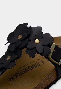 Κοντινό πλάνο του λουριού ενός σανδαλιού Birkenstock με τρία μαύρα δερμάτινα λουλούδια, καθένα με χρυσό κέντρο, και μια ρυθμιζόμενη μεταλλική αγκράφα.