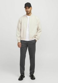 Bomber jacket beige claro, camiseta blanca, pantalones gris oscuro y zapatos negros. Los accesorios incluyen una gorra negra. Textura suave, diseño casual.