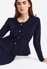Blazer de punto azul marino con cintura entallada, escote redondo y cinco botones metálicos. Textura suave con mangas largas y diseño de peplum.