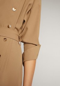 Gros plan sur une robe beige boutonnée avec une ceinture et une manche retroussée sur le bras d'une personne, sur un fond uni.