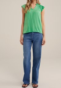 Zielona bluzka na krótki rękaw z dekoltem w serek i falbankami na ramionach, zestawiona z jasnoniebieskimi, szerokimi spodniami dżinsowymi oraz czarnymi klapkami.