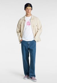 Beige jakke, hvid T-shirt med pink print, blå jeans og røde og hvide ternede slip-on sko, toppet med en sort beanie.