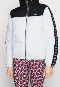 Puchowa kurtka z czarną górną częścią i białą dolną częścią, wyposażona w wysoki kołnierz, zamek błyskawiczny oraz czarne paski po bokach z logo.