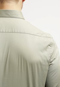 Grüne Button-Up-Bluse mit glatter Textur und taillierter Passform, ausgestattet mit einem schlichten Rückendesign und minimalistischen Nahtdetails.