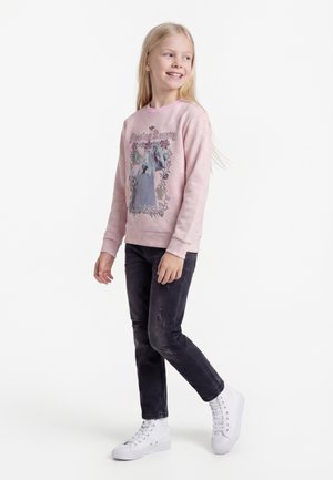 Smilende pige med langt blondt hår iført lyserød Tornerose-sweatshirt, sorte slidte jeans og hvide højtstående sneakers, står sidevej.