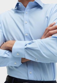 Jasnoniebieska koszula z długim rękawem i kołnierzykiem w kształcie spiczastym, z białymi guzikami. Tkanina wydaje się gładka i lekka.