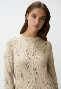 Beige gestrickter Pullover mit einem strukturierten Zopfmuster und offen gearbeiteten Details. Rundhalsausschnitt, lange Ärmel und weicher Stoff.