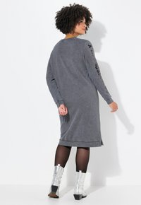Robe grise à manches longues avec une coupe décontractée, présentant des poignets côtelés, un ourlet arrondi et des lettres noires le long des manches. Associée à des bottes argentées.