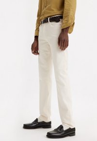 Pantalones de algodón blancos con un corte clásico, combinados con un cinturón de cuero trenzado marrón y mocasines de cuero negros. Textura suave, diseño de pierna recta.