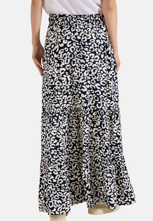 Persona que lleva una falda maxi con estampado de leopardo en blanco y negro con cintura elástica y zapatillas blancas, de pie contra un fondo liso.