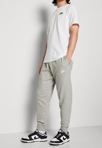 Persona vistiendo una camiseta blanca de Nike, pantalones de chándal grises de Nike y zapatillas Nike en blanco y negro, de pie con las manos en los bolsillos.