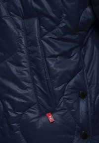Marineblauwe gewatteerde jas met een glanzende afwerking, voorzien van een rode label, zwarte knopen en een gestructureerd ruitpatroon op de stof.
