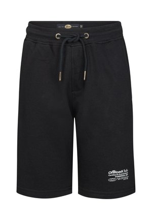Jongen met krullend haar staand, draagt een wit T-shirt, zwarte korte broek, witte sokken en witte sneakers tegen een effen lichte achtergrond.
