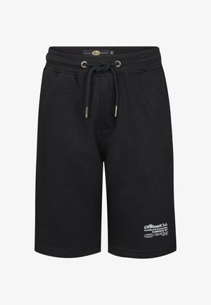 Jongen met krullend haar staand, draagt een wit T-shirt, zwarte korte broek, witte sokken en witte sneakers tegen een effen lichte achtergrond.