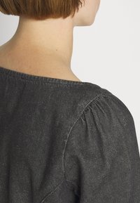 Cheveux courts bruns et nuque nue d'une personne portant un haut en tissu texturé gris foncé vue de dos sur fond blanc.