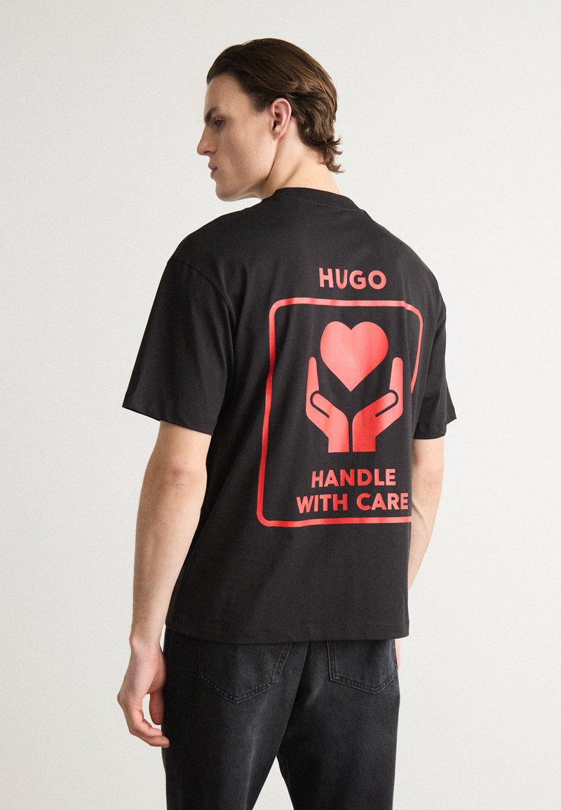 Juodas medvilninis marškinėlis su dideliu raudonu grafiniu piešiniu ant nugaros, vaizduojančiu rankas ir širdį, žymėtu "HUGO HANDLE WITH CARE."