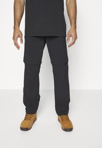 Zwarte, lichte cargobroek met ritsvakken, gedragen met tan wandelschoenen. De broek heeft een omkeerbaar ontwerp voor een verstelbare lengte.