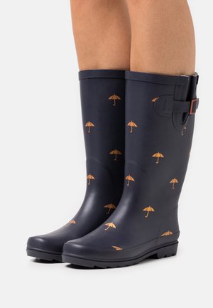 Bottes de pluie bleu marine montant jusqu'au genou avec motifs de parapluies orange, munies de sangles à boucles sur le côté et semelles texturées sur des jambes à peau claire.