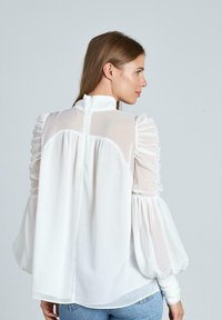 Blusa blanca con mangas abullonadas y detalles con volantes. Tejido transparente con un cuello alto y un cierre vertical en la parte posterior. Textura suave, corte relajado.