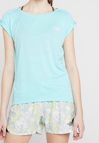 T-shirt de sport bleu clair à manches courtes avec un col rond et un logo. Associé à un short gris avec un motif abstrait coloré.