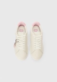 Κρεμ χρώματος αθλητικά παπούτσια με ροζ εσωτερική επένδυση και σόλες σε σχήμα καρδιάς, το δεξί παπούτσι διακοσμημένο με ένα ασημένιο μενταγιόν.