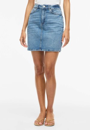 Frau trägt einen knielangen blauen Jeansrock mit vorderer Naht, Taschen und beigefarbenen Zehensandalen vor weißem Hintergrund.