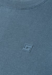 Gros plan d’un pull tricoté bleu avec un col côtelé et un petit logo géométrique en relief au centre.