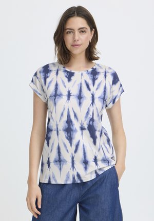 Nainen, joka pukeutuu lyhythihaiseen valkoiseen T-paitaan, jossa on sininen liukuvärjäyskuvio, ja sinisiin housuihin. Hän seisoo käsi taskussa yksiväristä taustaa vasten.