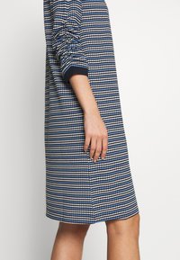 Robe marine avec un motif géométrique en bleu et blanc. Elle possède des manches longues, une coupe décontractée et une coupe droite arrivant à mi-cuisse.