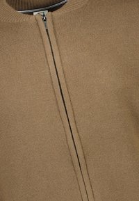 Brun stickad jacka med hög krage, med en framdragkedja och kontrasterande svart sömnad. Materialet är mjukt och texturerat.