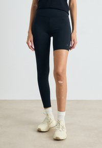 Sorte tettsittende sykkelshorts med en glatt tekstur og høy midje, parret med kremfargede joggesko og hvite sokker. Minimal merkevare synlig.