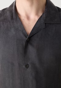 Camisa de botón gris con una tela ligeramente texturizada, que presenta un cuello con muesca y un detalle de un solo botón. Vista cercana del área del pecho.