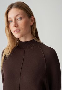 Bruine trui met een hoge, geribbelde kraag, een gestructureerd breisel en raglanmouwen. Heeft een centrale naden detail.