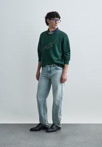 Πράσινο φούτερ με κεντημένο κείμενο, φορεμένο πάνω από μπλε πουκάμισο με γιακά. Το σύνολο ολοκληρώνουν ανοιχτόχρωμα τζιν και μαύρα δερμάτινα παπούτσια με κορδόνια.