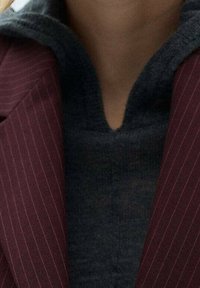 Personne portant un pull à fermeture éclair gris foncé sous un blazer bordeaux à fines rayures.