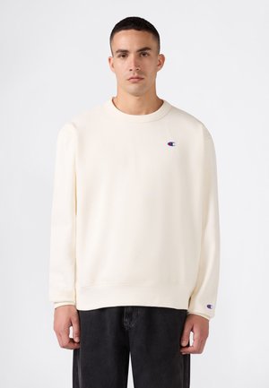 Mann trägt schlichten off-white Pullover mit kleinen Champion-Logos auf Brust und Ärmel, kombiniert mit schwarzen Hosen, steht vor weißem Hintergrund.