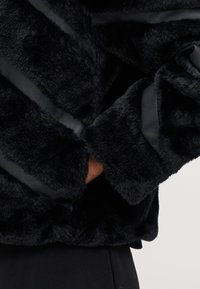 Μαύρο τζάκετ από τεχνητή γούνα με πλούσια υφή και διαγώνιες σατινέ ρίγες. Διαθέτει στρογγυλή ραφή στο κάτω μέρος και φαρδιές μανσέτες, προσφέροντας μια άνετη εμφάνιση.